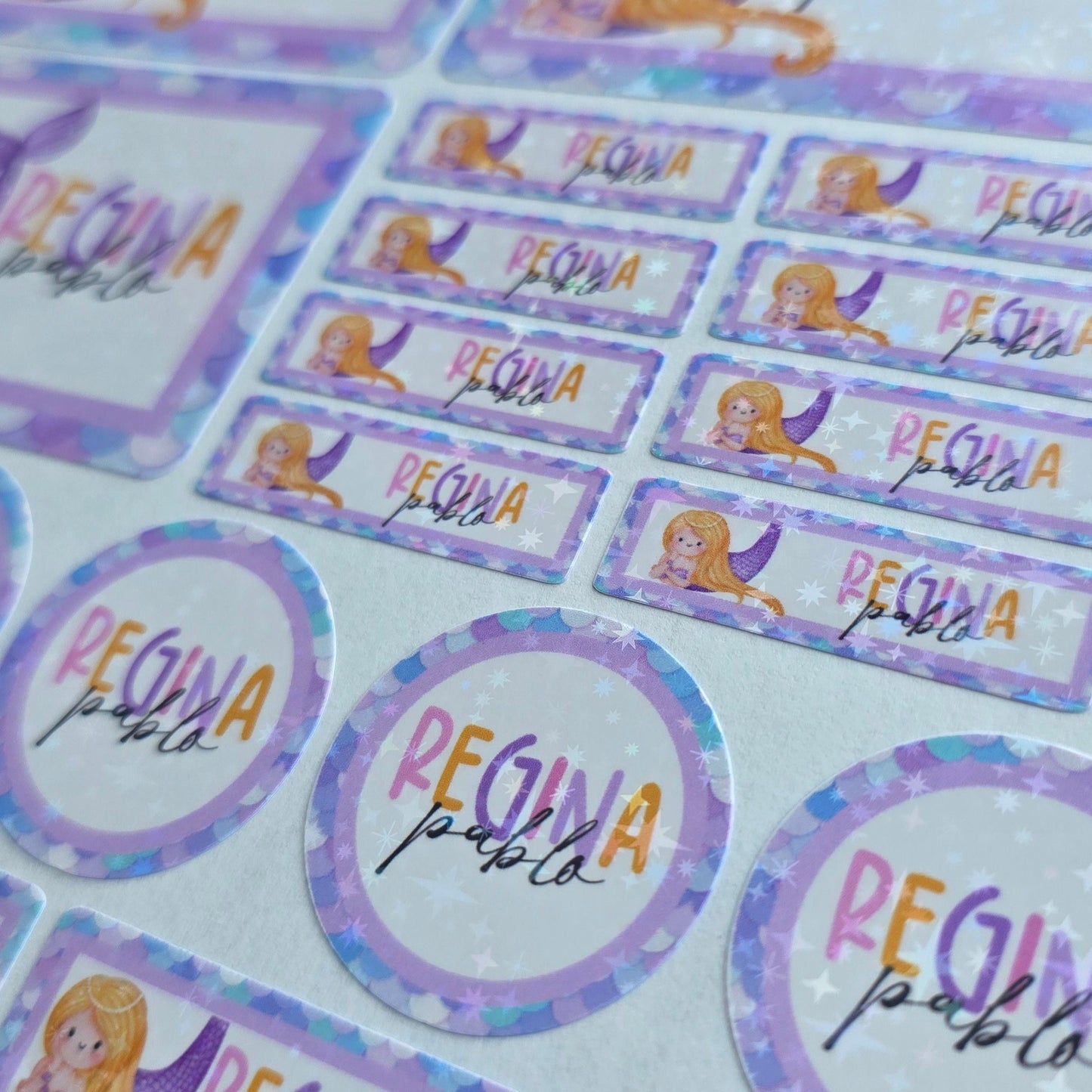 Close-up of personalized holographic mermaid labels with 'REGINA' and 'pablo' for school supplies.
Custom die-cut mermaid name stickers featuring iridescent scales & a cartoon mermaid, perfect for kids' gear.
Vibrant personalized mermaid-themed school labels with rounded rectangular and circular shapes, names visible.